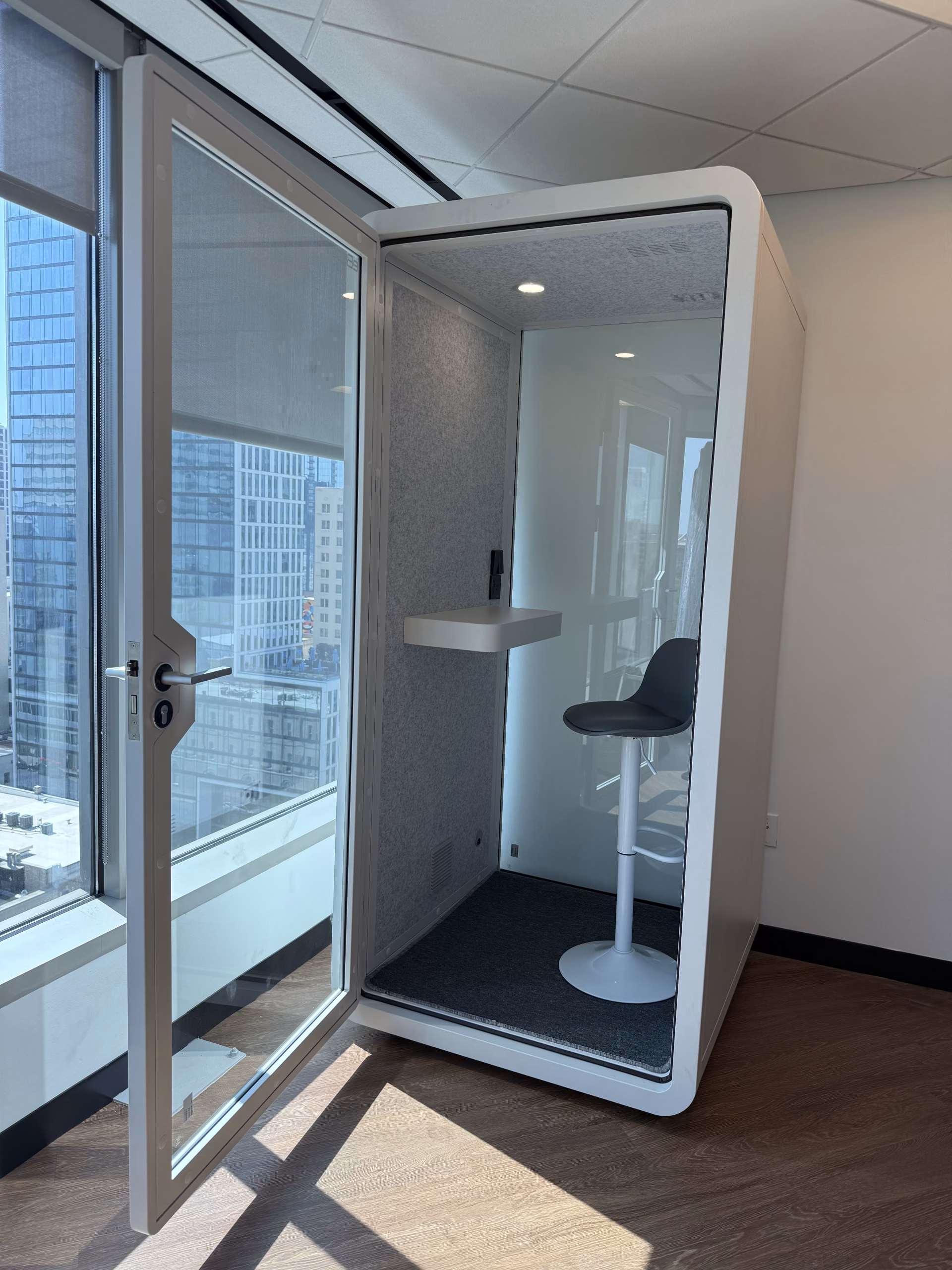 A single-occupancy acoustic phone booth installed against a high-rise window wall, door open on the fitted stool and shelf.