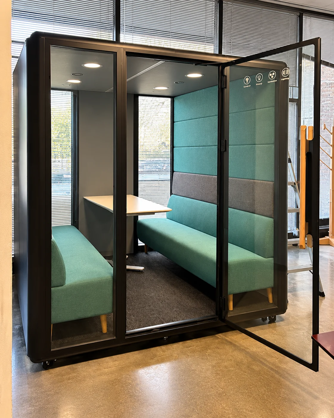 A glass-walled acoustic meeting pod with upholstered bench seating, installed on an office floor.