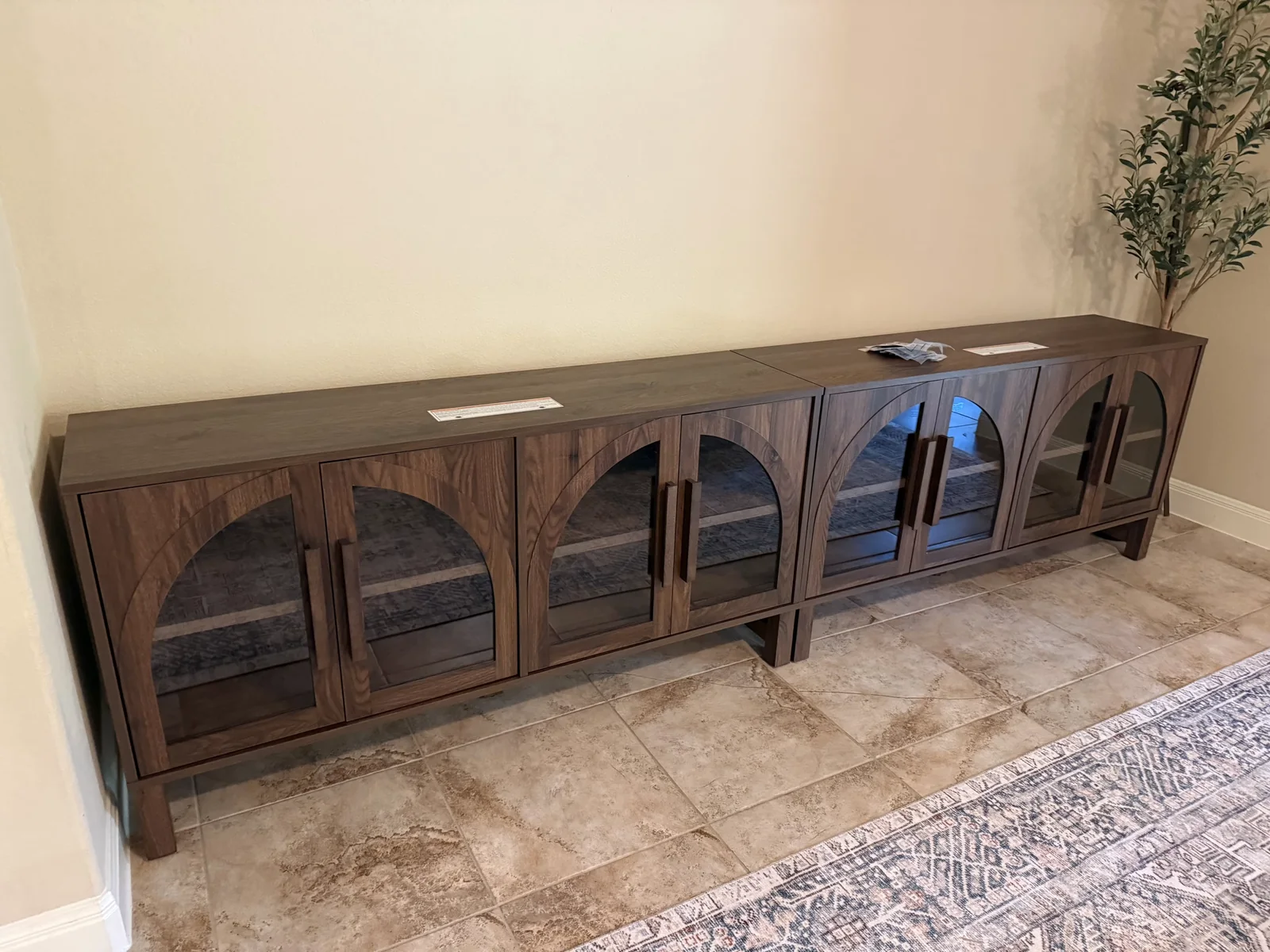 A matched pair of walnut credenzas with arched glass doors, assembled and set end to end against the wall of a finished room.