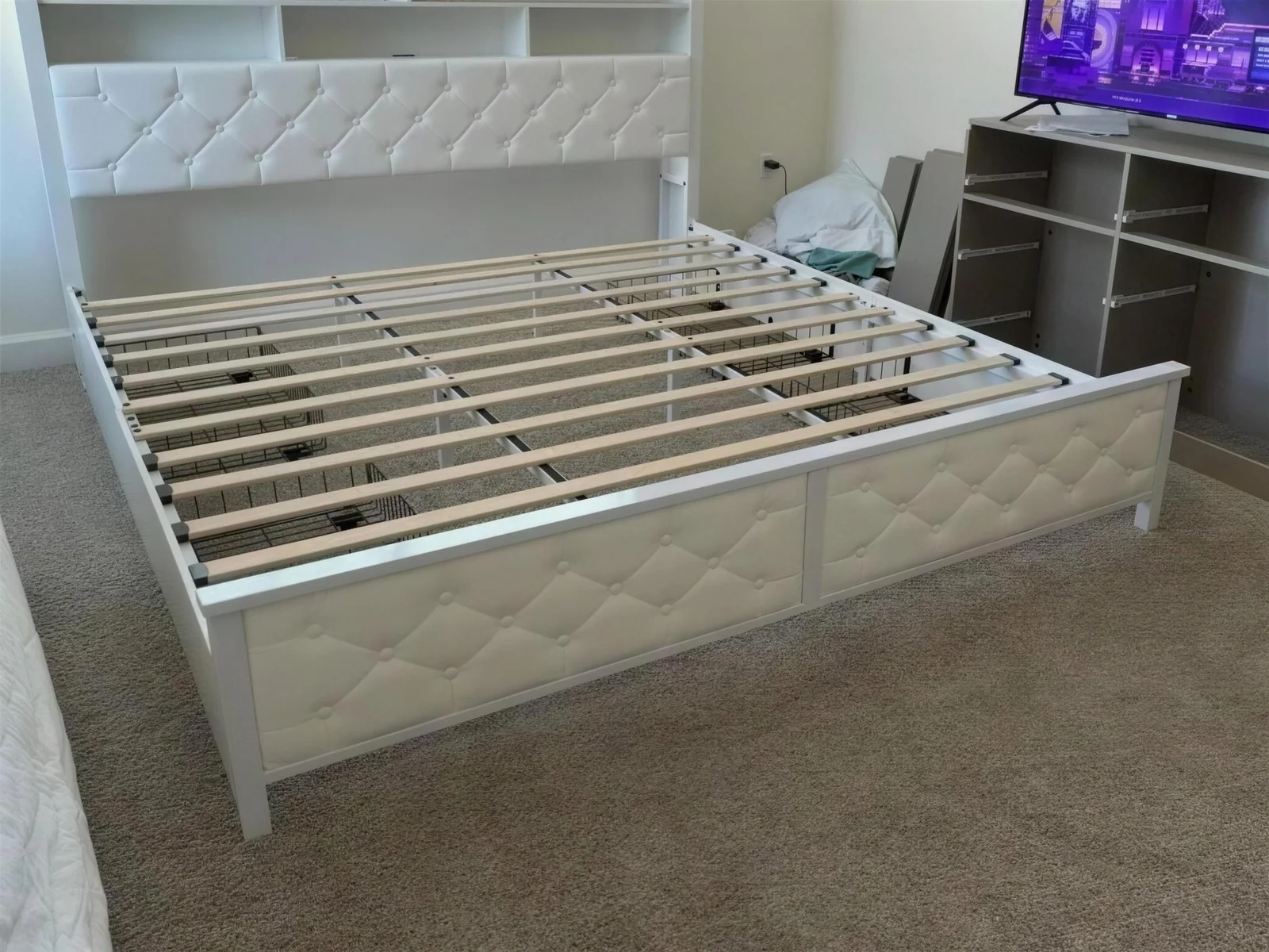 A white upholstered bed frame assembled and slatted in a unit between residents, with a part-built storage unit still in flat panels behind it.